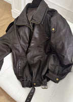 Women’s dark brown leather bomber jacket laid flat, zip front, snap pockets, belt strap