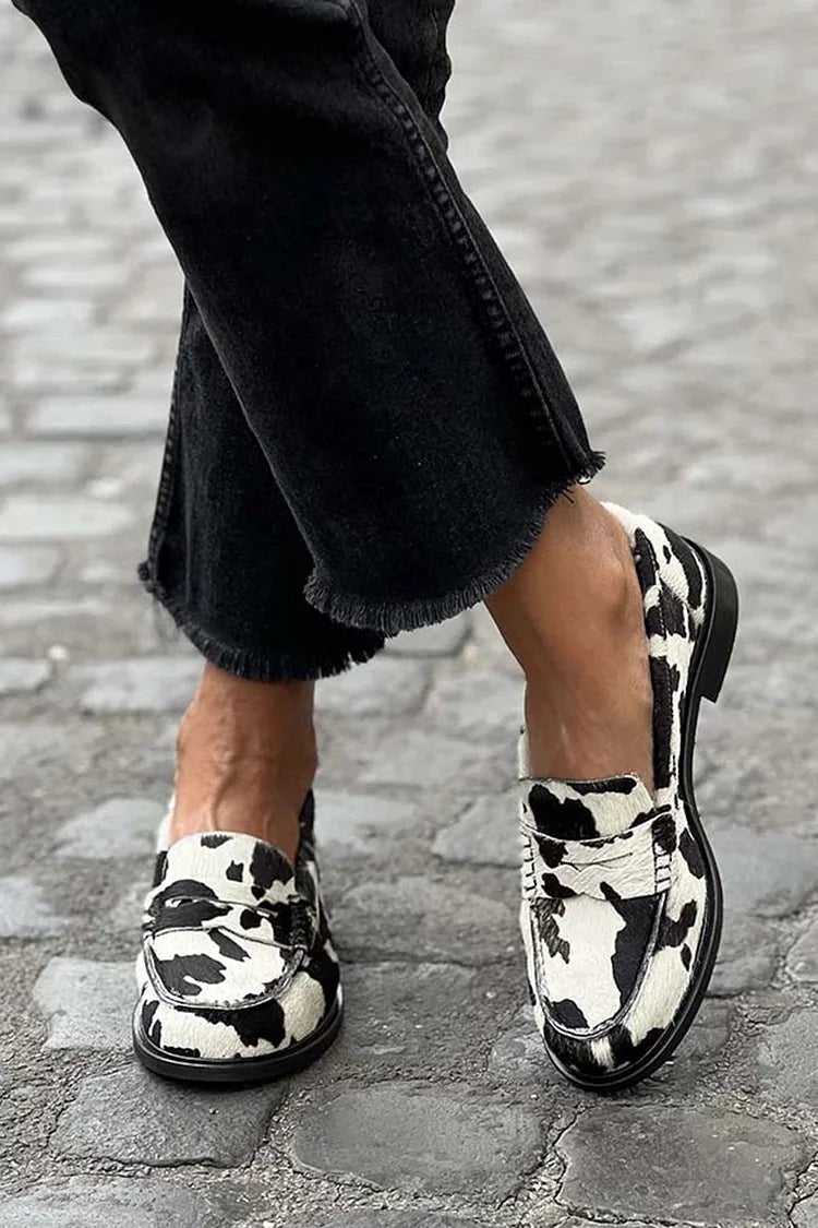 Women's cow print slip-on loafers in black and white, worn with black pants.
